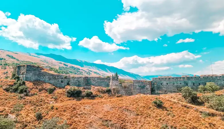 Stone walls of Gjirokastër Castle stretch across a golden hillside, backed by rolling green mountains under a bright blue sky with scattered clouds.
