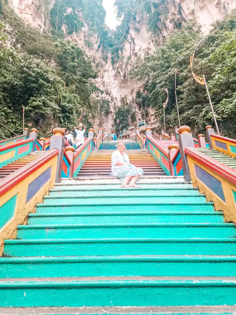 Em sitting on the colourful rainbow steps leading up to Batu Caves, surrounded by lush jungle and dramatic limestone cliffs, a highlight on many 2 week Malaysia itineraries.