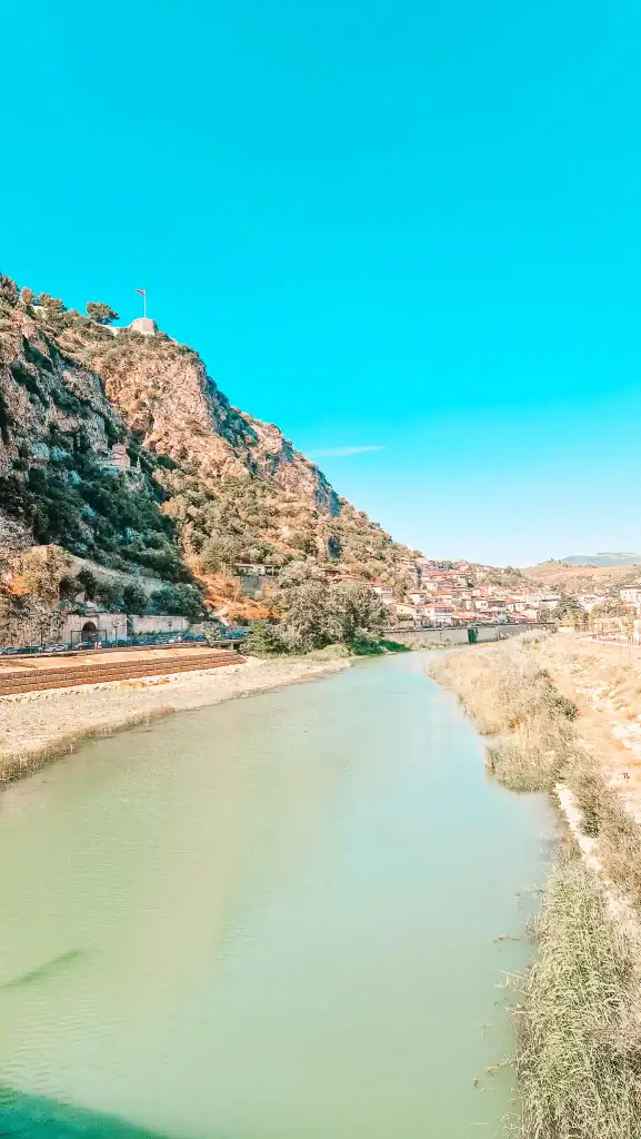 A tranquil river curves alongside the traditional white houses of Berat, with a rocky cliff and fortress flying the Albanian flag above. A scenic stop on a 10 day Albania itinerary.