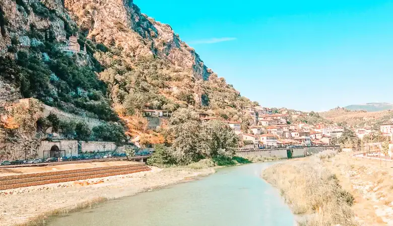 A turquoise river winds through a sunlit valley below a rugged hill with a small stone structure and flag on top, with the town of Berat nestled in the distance under a vivid blue sky.