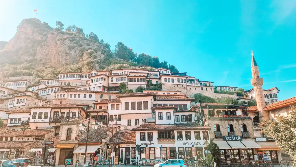 Cluster of Ottoman-style homes with white façades and tiled roofs climbing up the hill in Berat’s historic Mangalem Quarter, with a mosque minaret and Albanian flag atop the rocky peak.