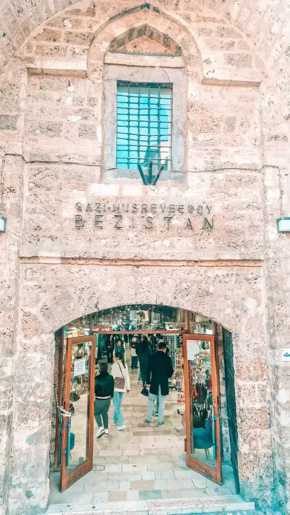 Stone archway entrance to Gazi Husrev-begov Bezistan, a covered market in Sarajevo, with people walking inside past souvenir stalls. The historic architecture frames a bustling glimpse into traditional Bosnian commerce.