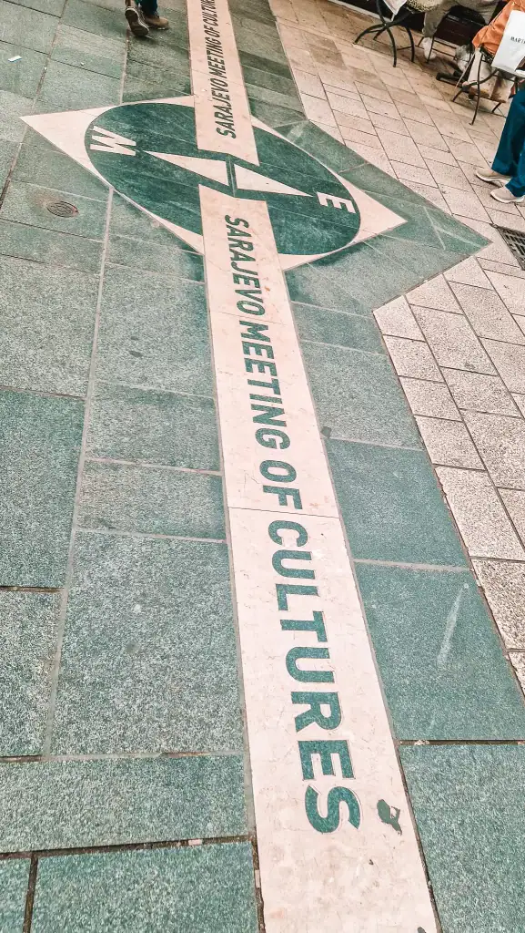 Green and white directional marker on a tiled pedestrian street in Sarajevo reading “Sarajevo Meeting of Cultures,” symbolising the city’s blend of Eastern and Western influences. A pair of feet visible in the corner shows the marker’s street-level position.