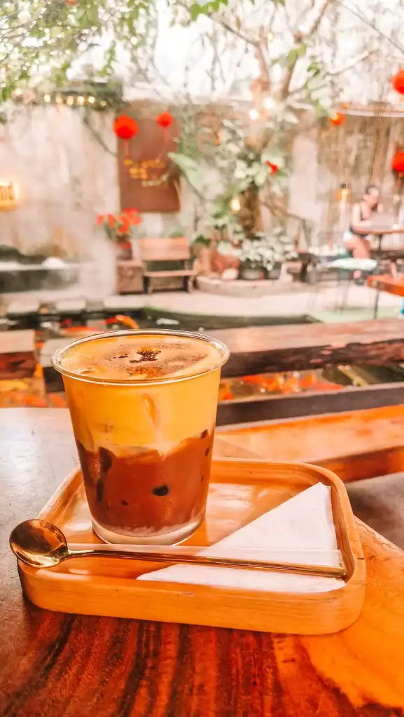 A creamy Vietnamese egg coffee served in a glass over ice, set on a wooden tray in a peaceful garden café with koi pond and red lanterns in the background.