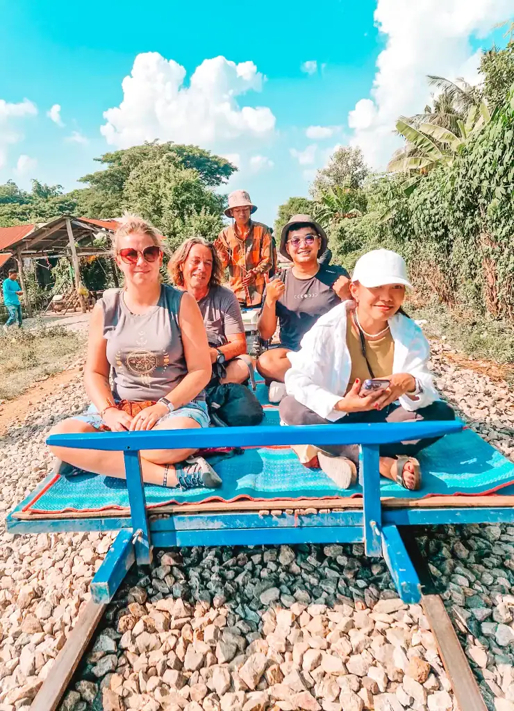 Em and a group of travellers sitting on the colourful bamboo train in Battambang, Cambodia, with bright skies and lush countryside passing by in the background.