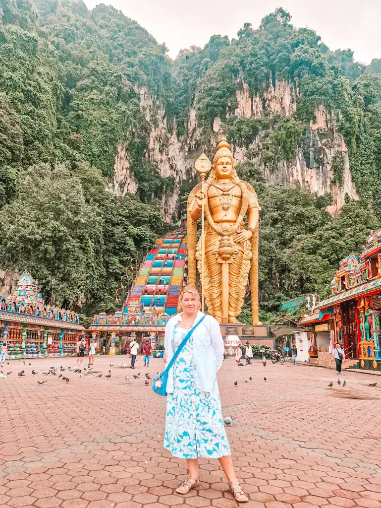 Em standing at the base of the towering golden Lord Murugan statue in front of Batu Caves, with the vividly painted staircase and forested limestone cliffs in the background.