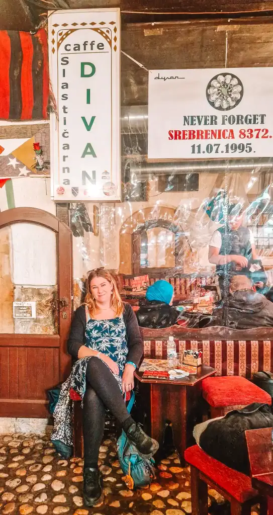 Em sits at a table inside Caffe Divan in Sarajevo, surrounded by traditional decor and a banner memorialising the Srebrenica massacre. The scene conveys the deep cultural and historical layers found in Sarajevo’s cafés.
