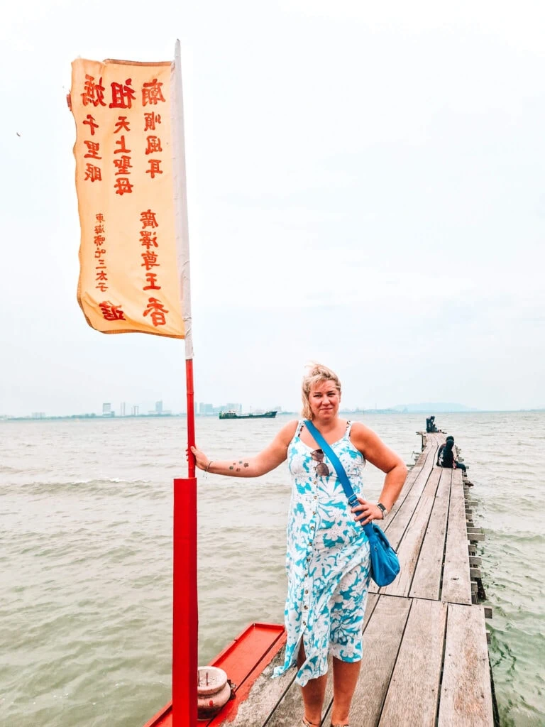 Em posing on a wooden jetty overlooking the sea in Penang, holding a red flagpole with Chinese script, with the city skyline faintly visible in the distance.