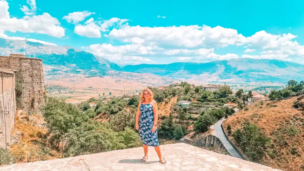 Em stands confidently on a stone castle platform, with terraced farmland, winding roads, and the blue mountains of Gjirokastër behind her.