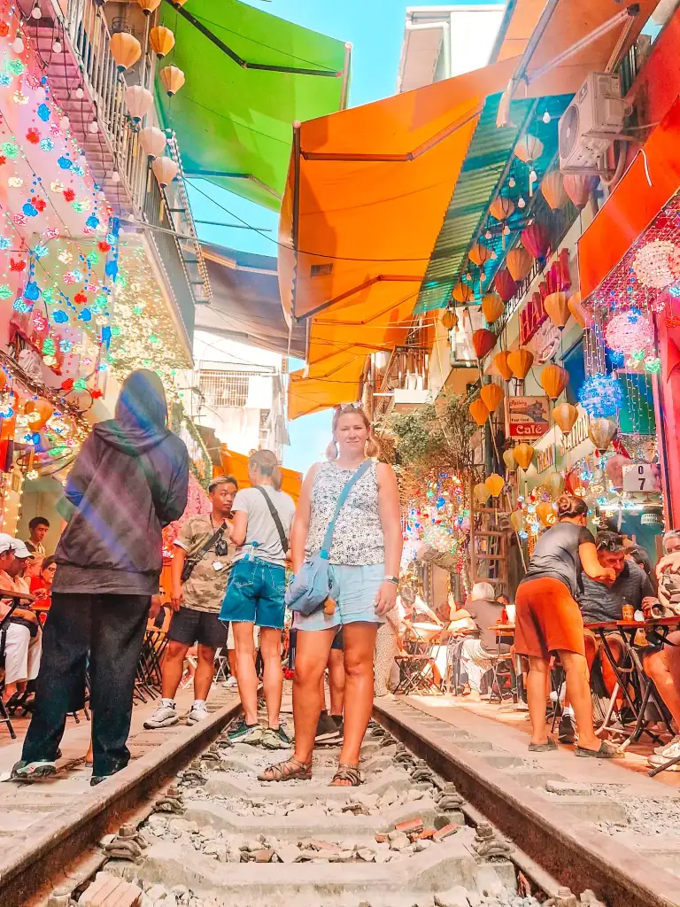 Em stands on the railway tracks of Hanoi’s famous Train Street, surrounded by cafés, hanging lanterns, and bustling tourists under colourful awnings.