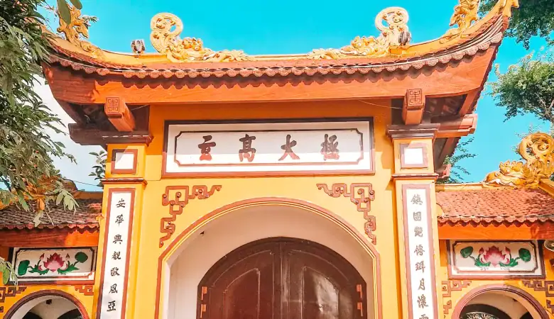 Em stands in front of a bright yellow and red temple gate with ornate rooftop dragons and vertical Vietnamese calligraphy on pillars, under a clear blue sky.