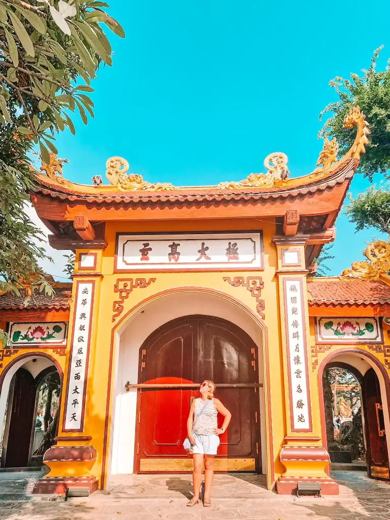 Em stands in front of a bright yellow and red temple gate with ornate rooftop dragons and vertical Vietnamese calligraphy on pillars, under a clear blue sky.
