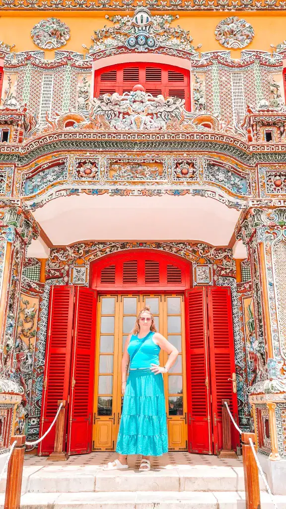 Em poses confidently in front of the vibrant, intricately decorated entrance of Khai Dinh Tomb in Hue, Vietnam, with detailed mosaics and bold red shutters behind her.