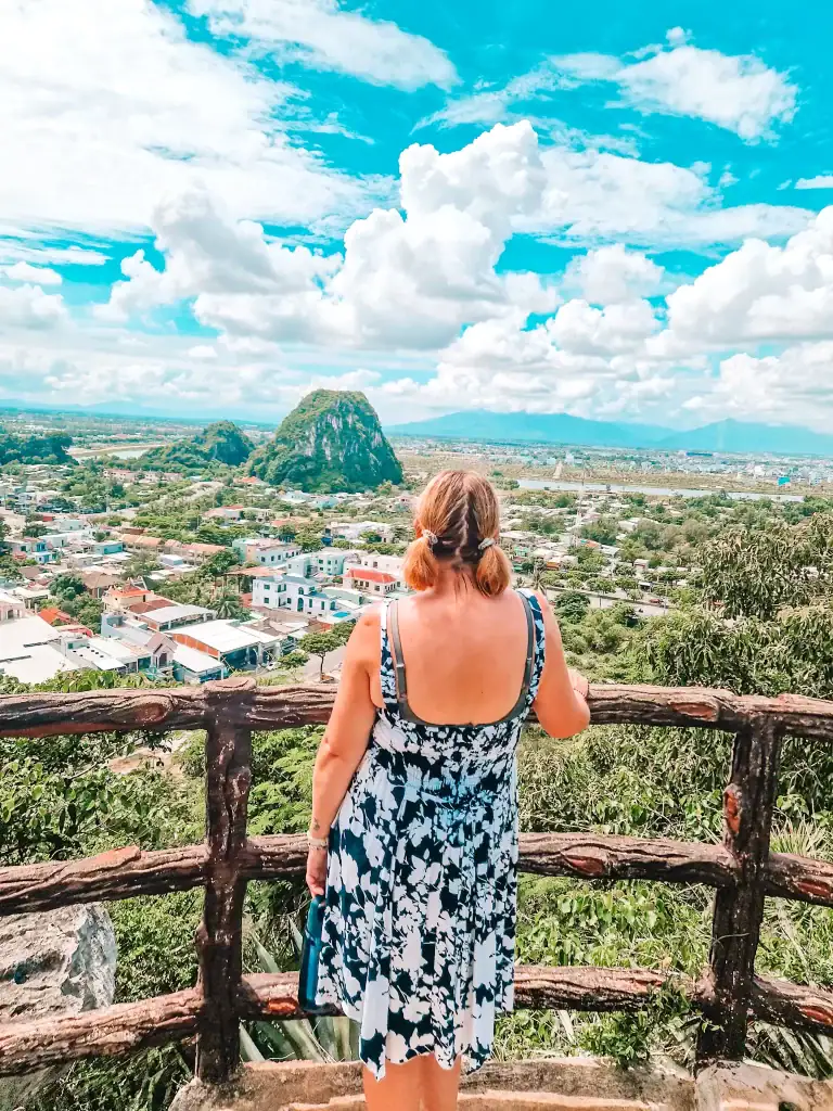 Em looks out over Da Nang city and the lush Marble Mountains from a wooden viewpoint, with a bright blue sky and scattered clouds above.