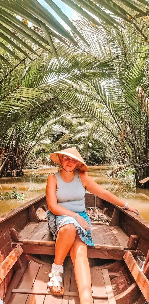 Wearing a traditional conical hat, Em relaxes in a small wooden boat gliding through a dense coconut palm forest in the Mekong Delta.