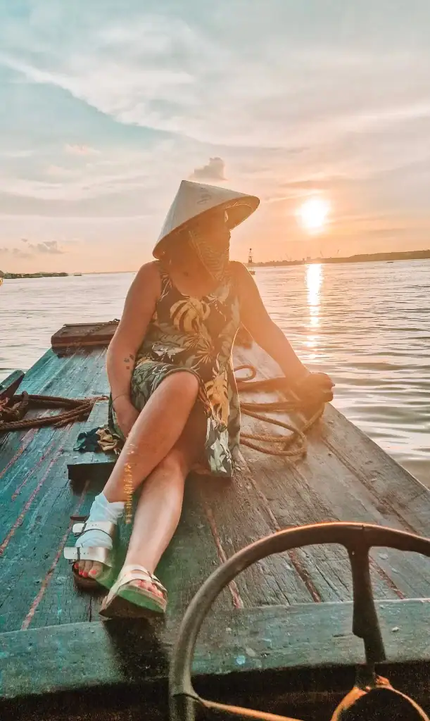 Em leans back on a wooden boat at sunset in the Mekong Delta, wearing a conical hat as the golden light reflects off the calm river water.