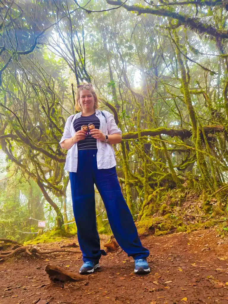 Em standing on a forest trail surrounded by moss-covered trees and mist, likely in the Mossy Forest of Cameron Highlands, holding two small figurines with a smile.