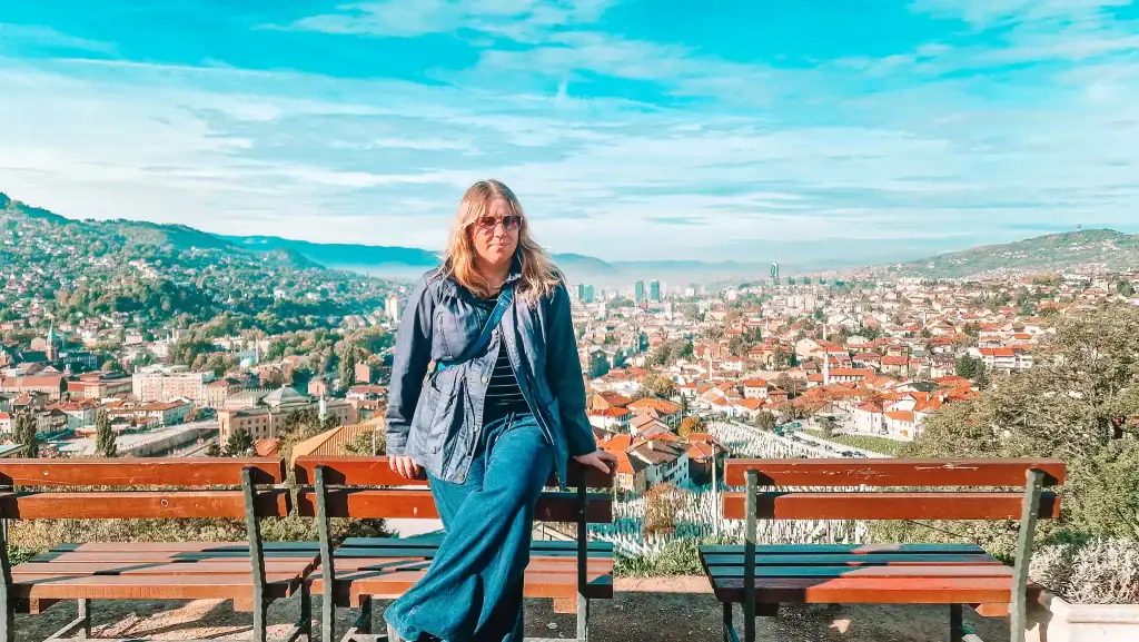 Em poses on a bench at the Yellow Fortress viewpoint, overlooking the sprawling cityscape of Sarajevo with red-roofed houses and distant skyscrapers under a bright blue sky. A peaceful and scenic highlight among things to do in Sarajevo.