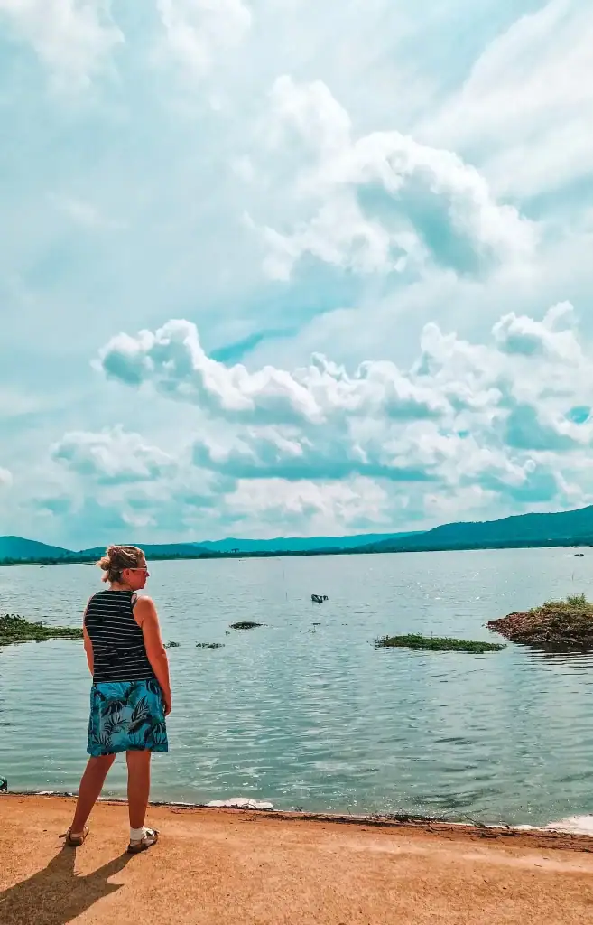 Em stands at the edge of a lake, gazing across the calm water with scattered floating vegetation and distant hills under a sky full of clouds.