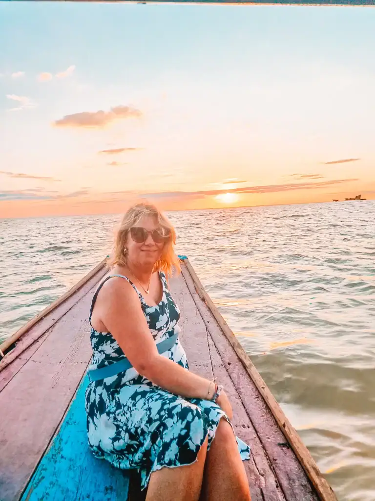 Em smiles at the camera while sitting on the bow of a long wooden boat at sunset on Tonlé Sap Lake, with gentle waves and warm golden light around her.