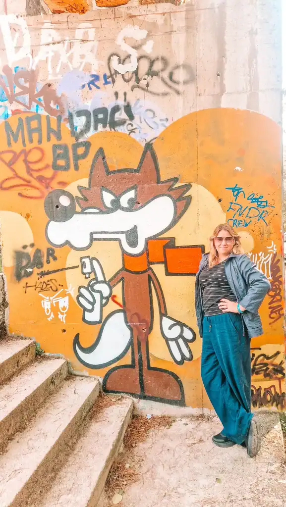 Em poses next to a cartoonish graffiti of Vučko, the 1984 Olympic Games mascot, painted on a yellow wall beside the bobsled stairs. The nostalgic artwork adds playful charm to this unique urban exploration site.