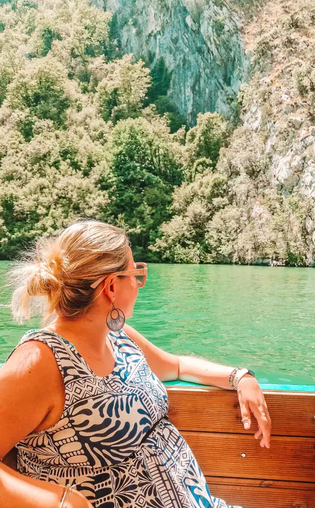 Em relaxes on a wooden boat cruising through emerald green waters, with forested cliffs rising steeply on the Shkodër side—a peaceful moment during her 10 day Albania itinerary.