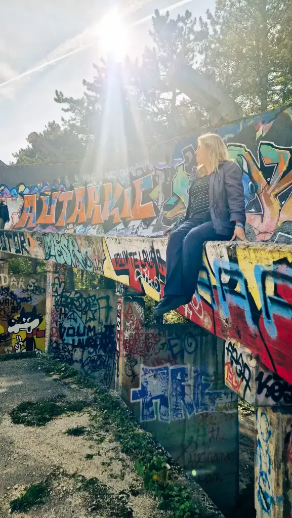 Em sits casually on the edge of the graffiti-covered Sarajevo bobsled track with sunlight streaming through trees. The scene captures both the urban decay and artistic revival of this Olympic-era site.