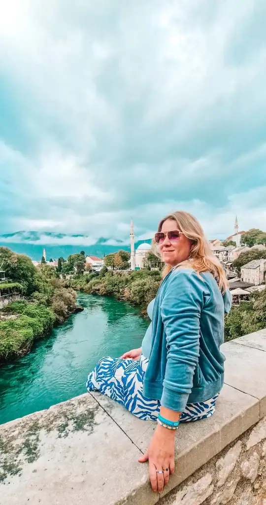 Em sits on the edge of a stone bridge in Mostar, overlooking the emerald Neretva River and mosque-filled skyline under moody skies. This peaceful moment captures the charm of exploring Mostar’s old town on foot.