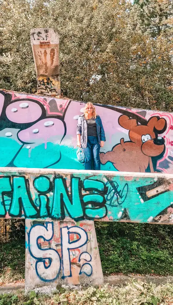 Em stands confidently on the graffiti-covered Sarajevo bobsled track beside a cartoon bear mural. The vivid street art pops against the natural backdrop, blending history and modern expression.
