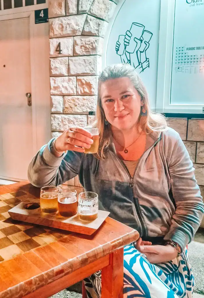 Em raises a glass during a beer tasting flight, seated outside at a wooden table with four small glasses of craft beer. Sampling local brews is a fun and relaxed option when looking for offbeat things to do in Mostar.