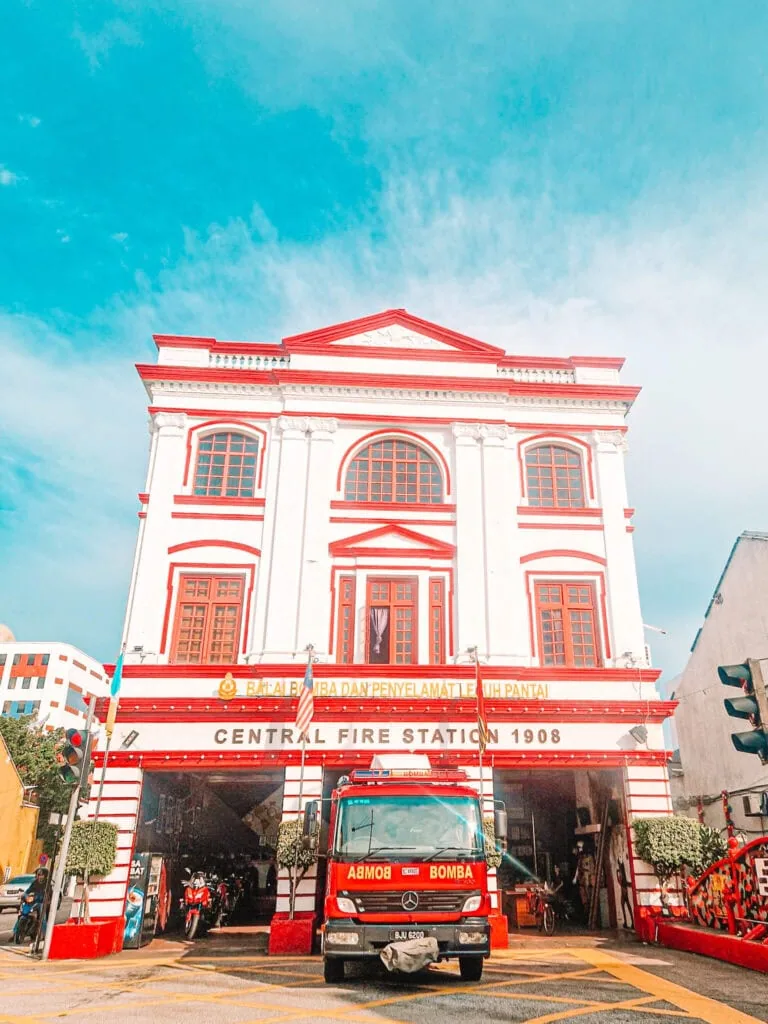 A bright red and white colonial-era fire station in George Town, Penang, with a fire engine parked outside under a sign reading “Central Fire Station 1908”.