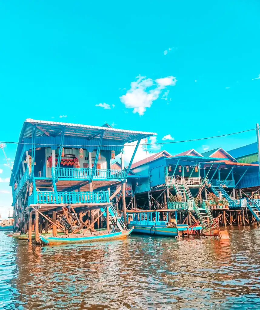 Traditional wooden stilt houses in bright blues and reds line the water in a floating village on Tonlé Sap Lake, with boats moored in front and laundry hanging from balconies.