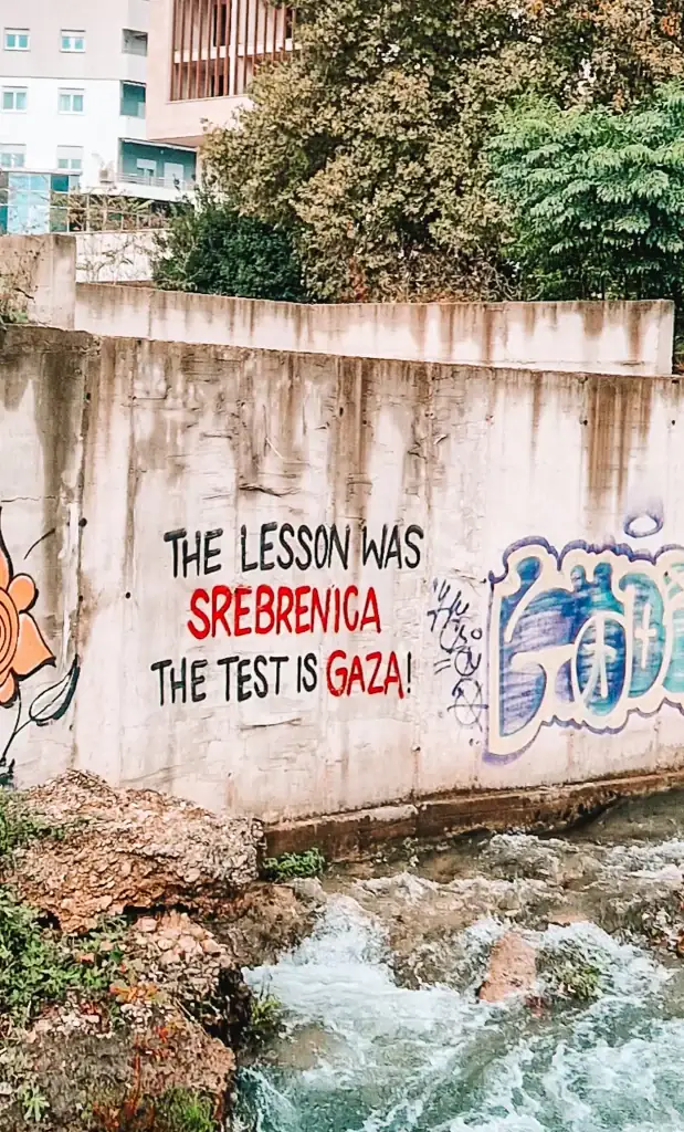 Graffiti on a concrete riverbank wall reads “THE LESSON WAS SREBRENICA. THE TEST IS GAZA!” in black and red letters. This politically charged street art in Mostar reflects the region’s deep awareness of global and historical conflict.