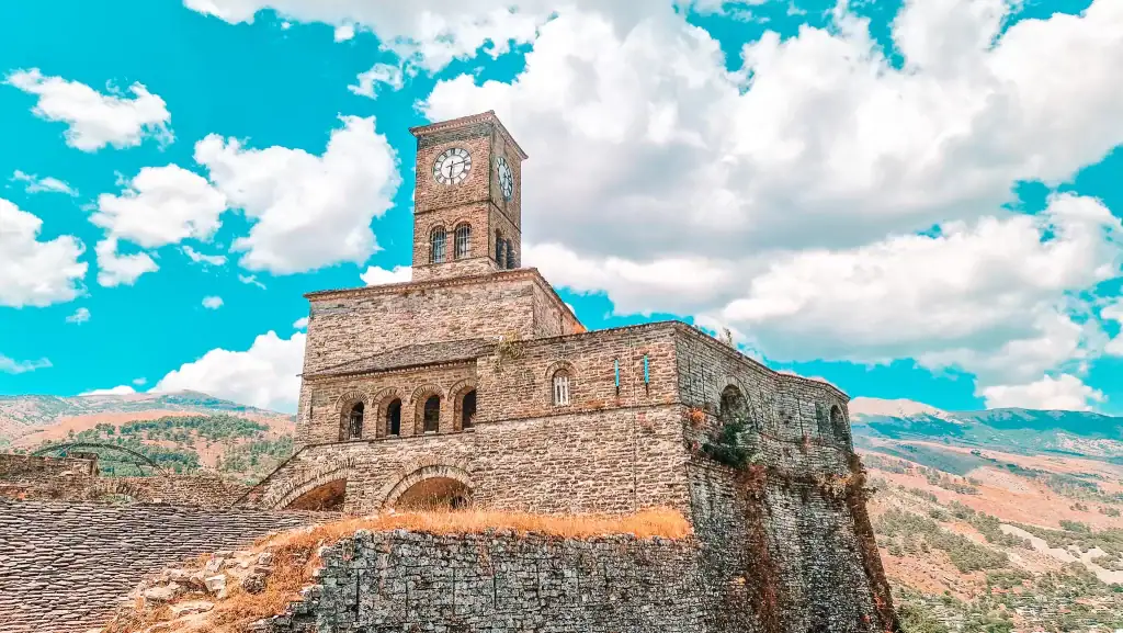 Gjirokastër Castle’s iconic clock tower rises above its stone base, framed by dramatic clouds and distant hills in the UNESCO-listed city.