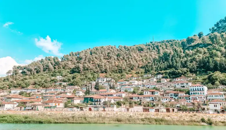 The colourful hillside quarter of Gorica in Berat is filled with traditional white homes and red roofs, lined up along the riverbank beneath a forested slope.