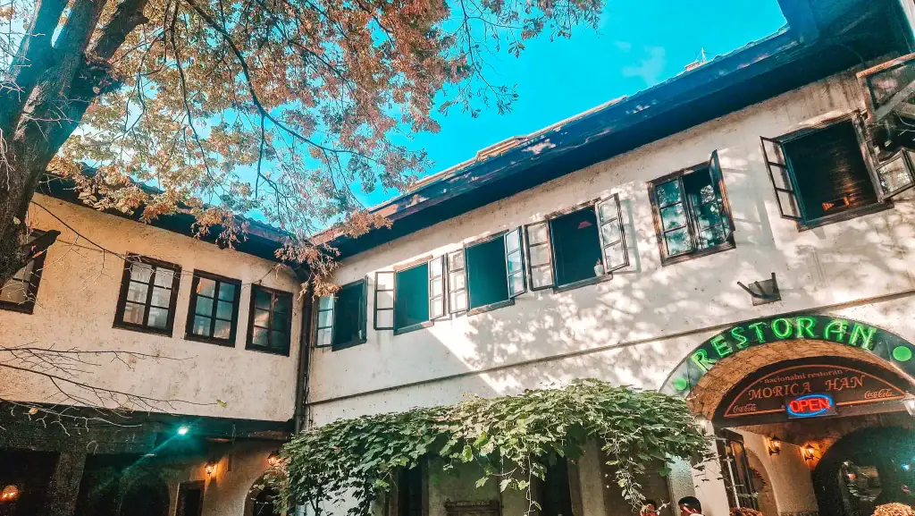 Courtyard view of the historic Morica Han in Sarajevo, with ivy-covered archways and open upper-floor windows. A neon “Restoran” sign marks the entrance to a traditional Bosnian restaurant.