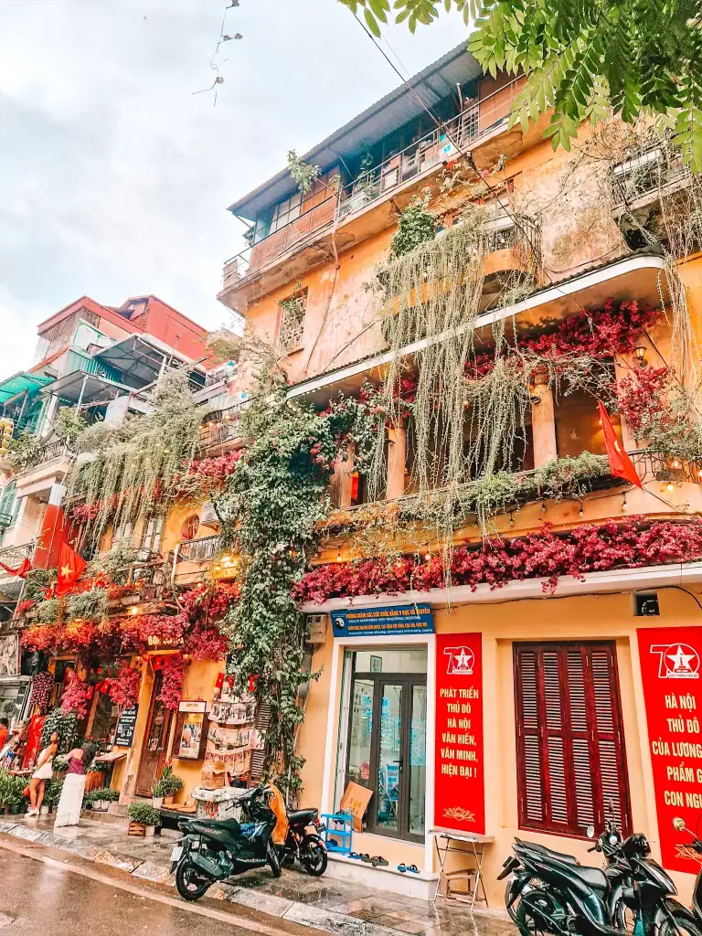 A colourful street in Hanoi, Vietnam, lined with motorbikes and covered in lush greenery and bougainvillaea, with shopfronts glowing warmly beneath colonial-style buildings.
