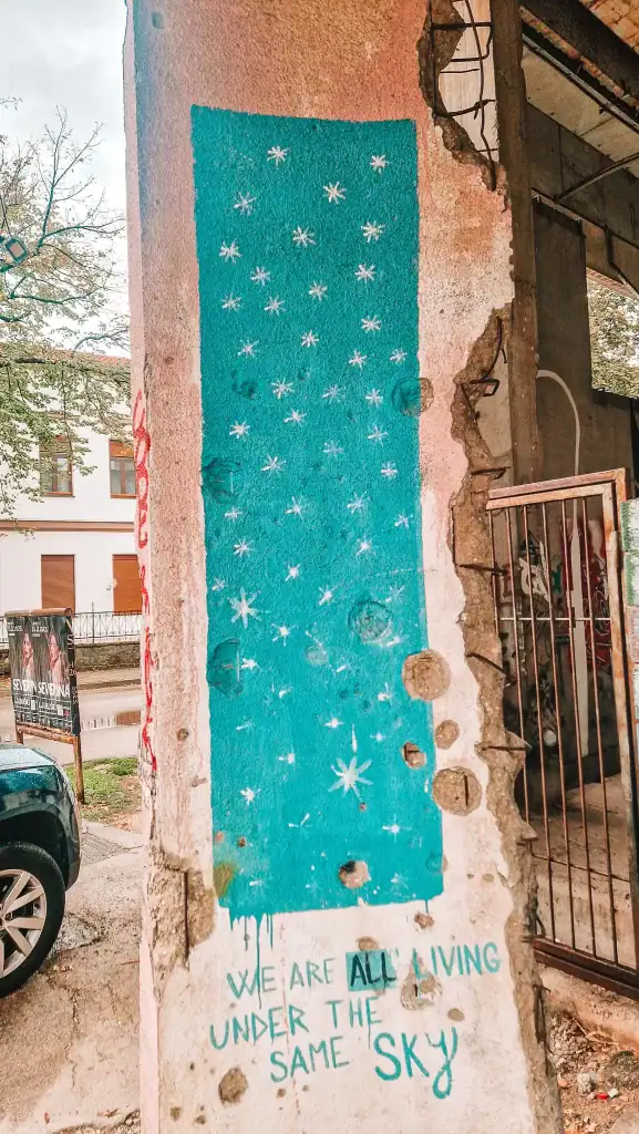 Blue mural painted on a damaged pink wall inside the former sniper tower, showing white stars and the phrase “WE ARE ALL LIVING UNDER THE SAME SKY.” This powerful urban artwork highlights Mostar’s post-war resilience.