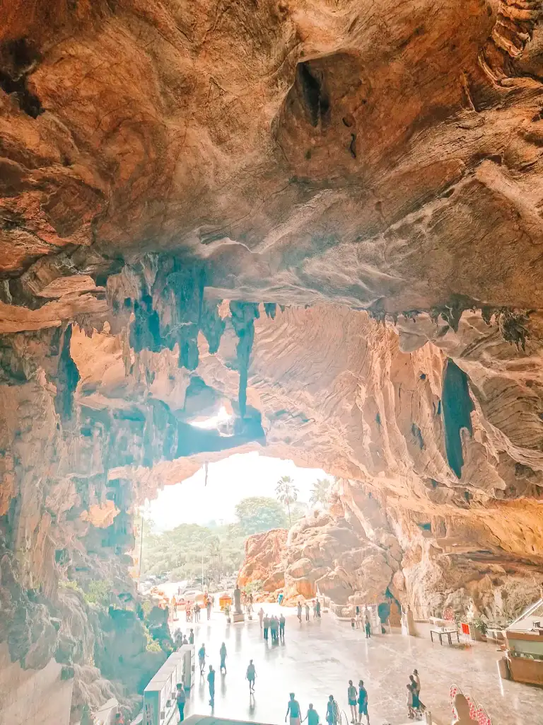 Looking out from inside a dramatic cave temple near Ipoh, with towering limestone walls, jagged stalactites, and visitors exploring the open courtyard bathed in soft natural light.