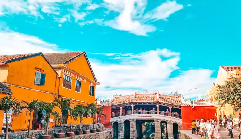 The iconic Japanese Covered Bridge in Hoi An, Vietnam, framed by bright yellow buildings and reflected in a narrow canal under a clear blue sky.