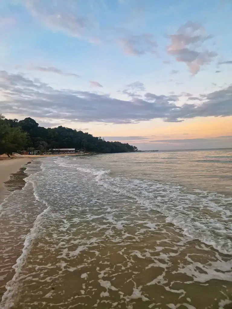 Gentle waves roll onto the soft sandy beach at Koh Rong Samloem during sunset, with pastel pink and blue clouds stretching across the sky and forested coastline in the distance.
