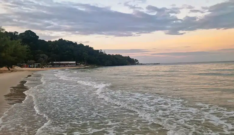 Gentle waves roll onto the soft sandy beach at Koh Rong Samloem during sunset, with pastel pink and blue clouds stretching across the sky and forested coastline in the distance.