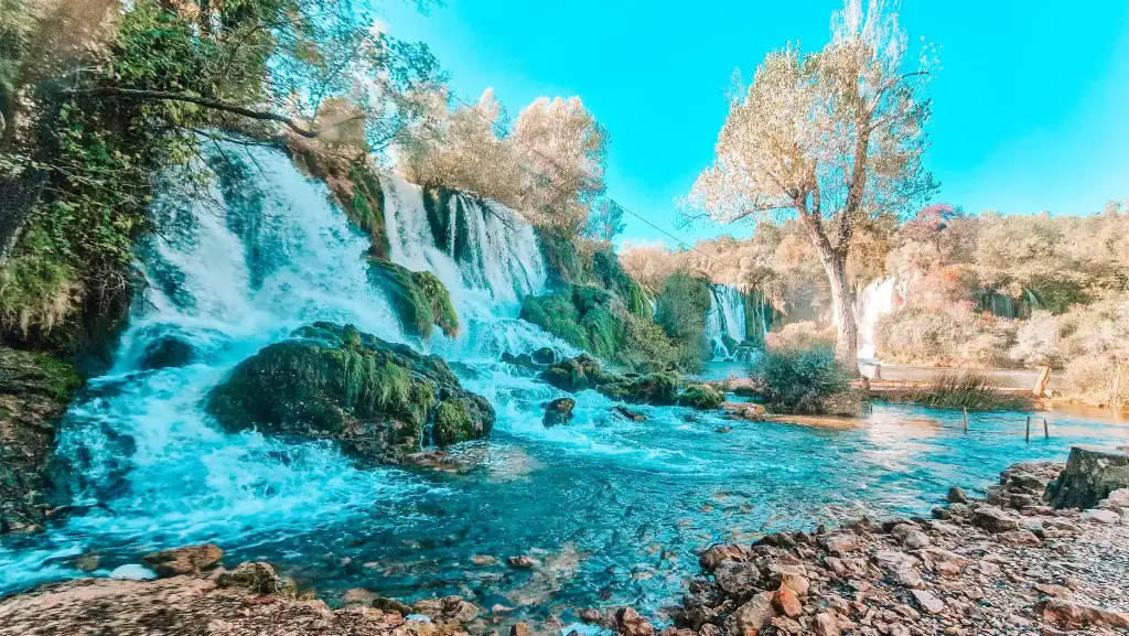 Multiple cascades of Kravice Waterfalls rush over moss-covered rocks into a blue-green river, surrounded by forest. A popular nature escape and one of the top day trips from Mostar.
