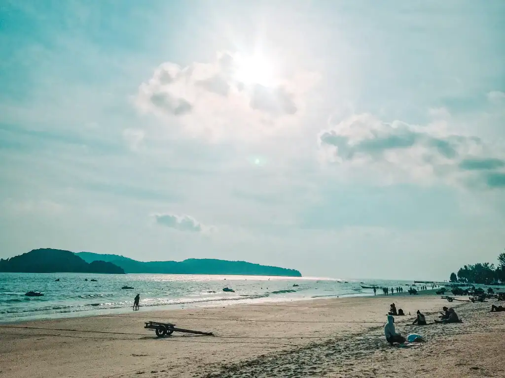 A calm sandy beach in Langkawi under soft sunlight, with a few people relaxing on the shore and distant islands visible across the shimmering sea.