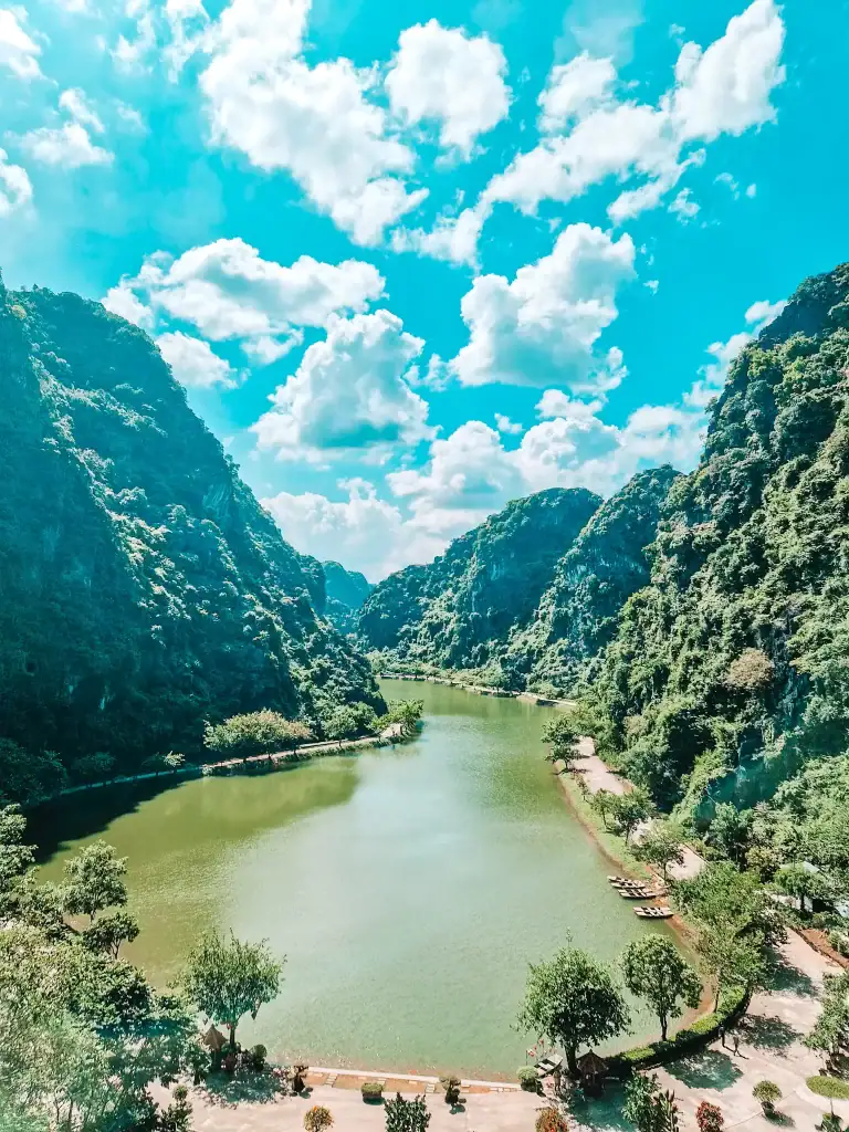 A sweeping view over a peaceful green river nestled between steep limestone cliffs in Ninh Binh, Vietnam, with scattered boats lining the water’s edge under a brilliant blue sky.