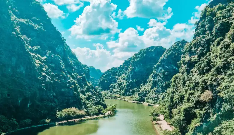 A sweeping view over a peaceful green river nestled between steep limestone cliffs in Ninh Binh, Vietnam, with scattered boats lining the water’s edge under a brilliant blue sky.