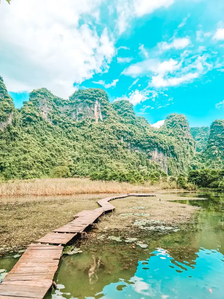A narrow wooden boardwalk winds over a still, lily-covered wetland in Ninh Binh, Vietnam, framed by towering, jungle-clad limestone karsts under a bright blue sky.