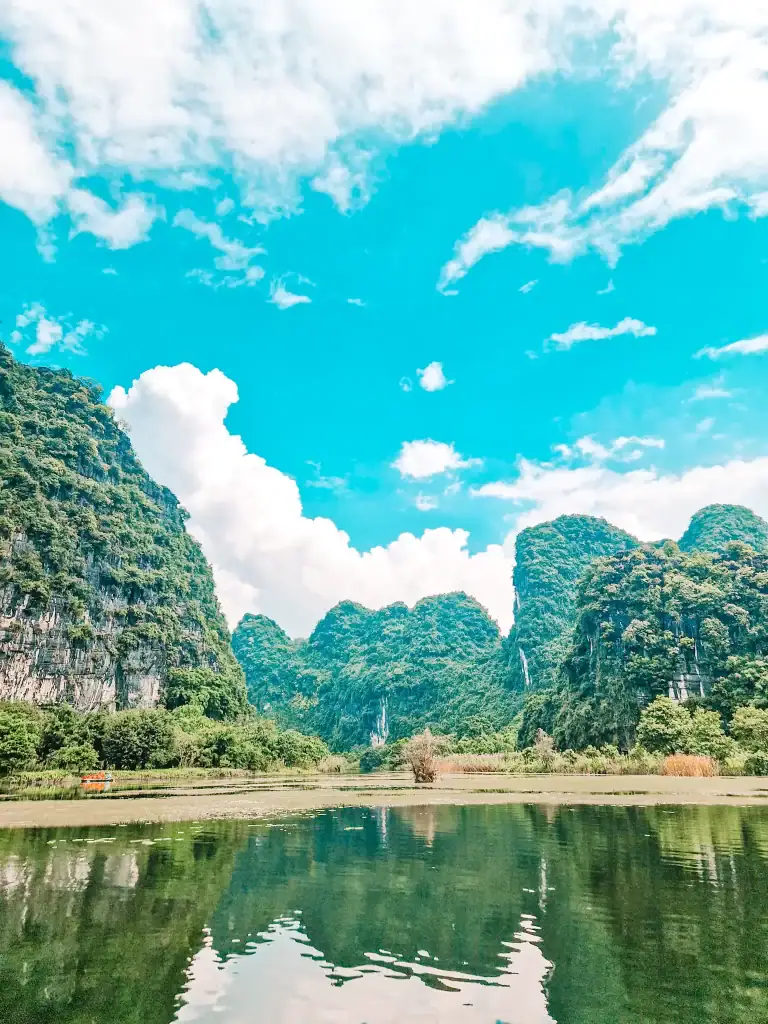 A glassy lake reflecting towering limestone karsts and lush greenery in Ninh Binh, Vietnam, under a sky dotted with soft white clouds.