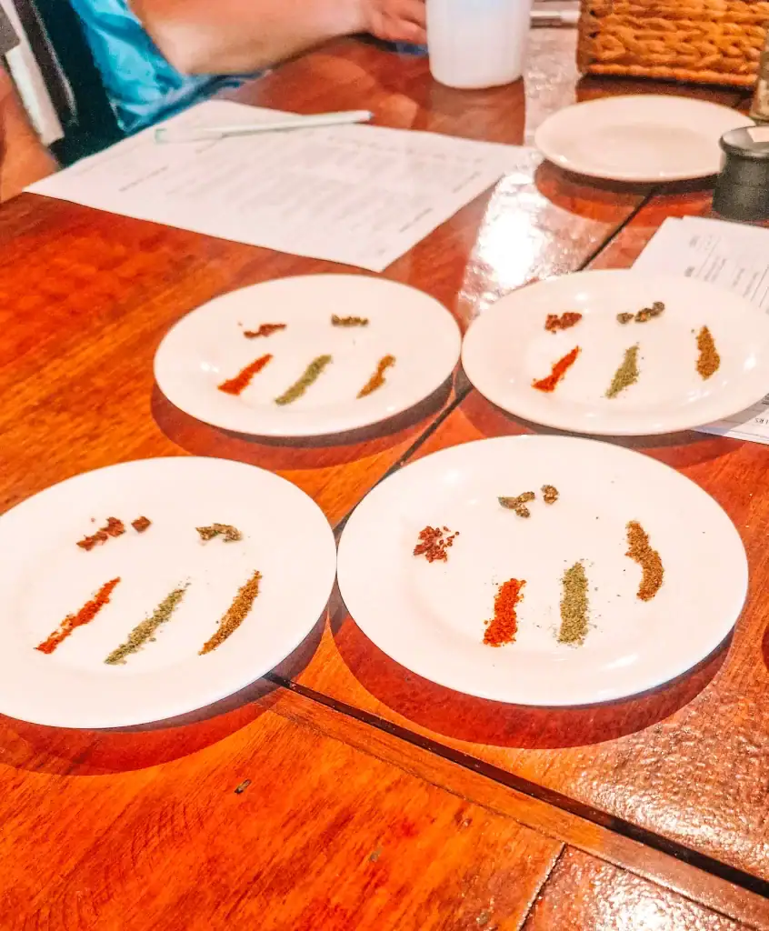 White tasting plates on a wooden table hold small piles of red, green, and black peppercorns during a Kampot pepper farm tour, with menus and a cup nearby.