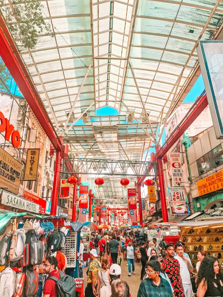 A bustling scene inside Kuala Lumpur’s Petaling Street Market, with red lanterns, hanging signs, and crowds of locals and tourists browsing through street stalls.